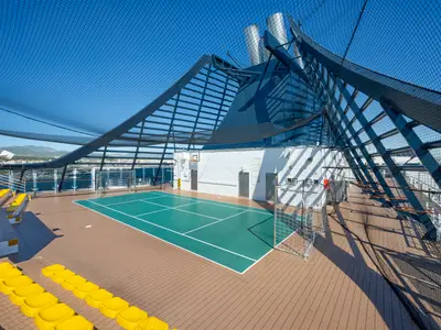 Blick auf den Multisportplatz an Deck der MSC Fantasia mit grüner Fläche für Tennis, Basketball und Fußball. Gelbe Tribünensitze und ein Schutznetz sind sichtbar.