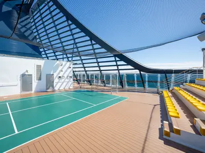 Ein Sportplatz auf dem Oberdeck der MSC Fantasia. Grünes Spielfeld, von einem Netz überdacht. Rechts gelbe Zuschauersitze, dahinter das weite Meer.