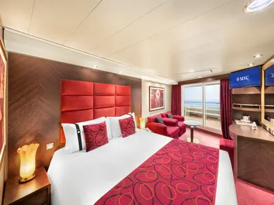 Blick in eine geräumige Suite an Bord der MSC Fantasia mit großem Doppelbett, rotem Kopfteil, Sofa und Balkon mit Meerblick. Der Raum ist elegant eingerichtet, mit Holzvertäfelung und einem Flachbildfernseher über einem Schminktisch.