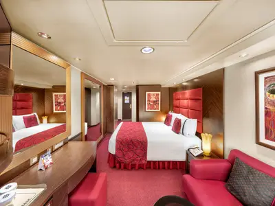 Geräumige Junior Suite an Bord der MSC Fantasia. Ein großes Doppelbett mit rotem Bettüberwurf und Kopfkissen, umgeben von dunklen Holz- und roten Polsterakzenten. Im Vordergrund ein Sofa und ein Schreibtisch mit Spiegel.