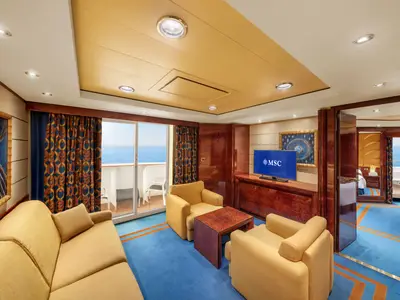 Geräumige Grand Suite Aurea an Bord der MSC Fantasia mit hellem Wohnbereich, Sofa, Sesseln, TV und privatem Balkon mit Meerblick. Ein Durchgang führt zum Schlafzimmer.