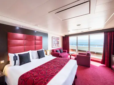 Eine geräumige Suite an Bord der MSC Fantasia in Rottönen mit großem Doppelbett, Sitzecke und Balkon mit weitem Meerblick.