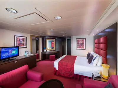 Geräumige Suite an Bord der MSC Fantasia. Ein grosses Doppelbett mit rotem Kopfteil, ein Sofa, TV und ein Schminktisch sind sichtbar. Warme Rottöne und dunkles Holz dominieren.