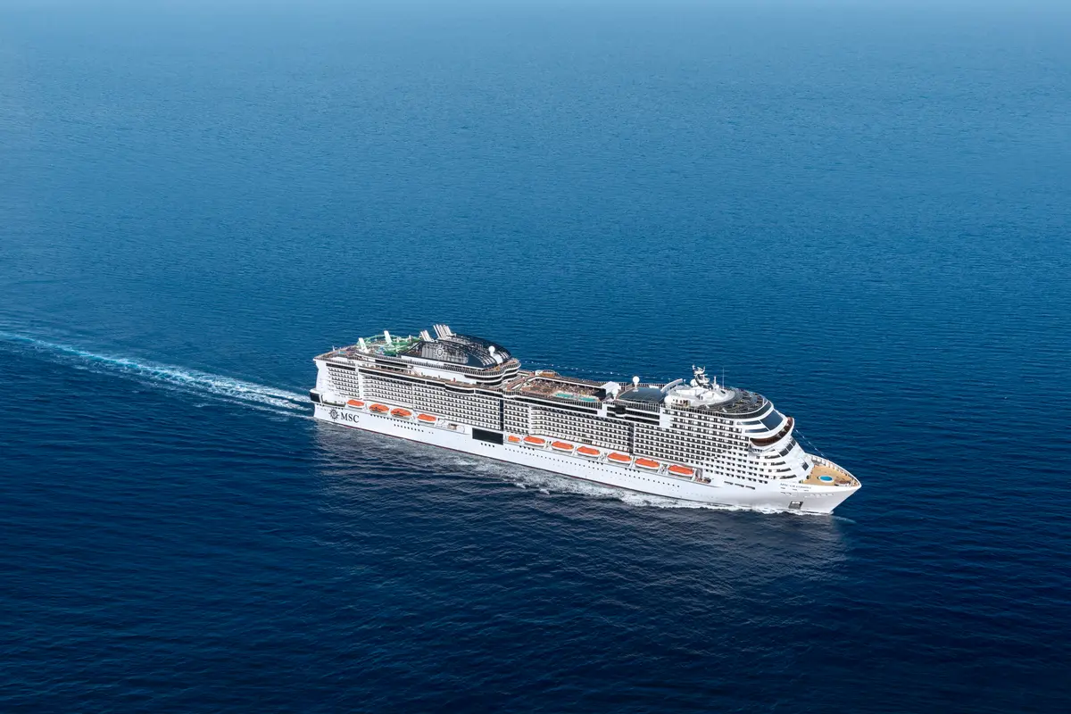 Vogelperspektive auf die MSC Grandiosa, die majestätisch auf tiefblauem Meer fährt und eine weiße Heckwelle zieht. Das moderne, weiße Schiff ist von vorne bis hinten sichtbar, mit vielen Balkonen und offenen Decks.