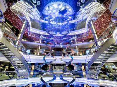Das futuristische Infinity Atrium der MSC Grandiosa. Zwei geschwungene Treppen und eine spiegelnde Kunstinstallation. Blaue Lichter schaffen eine moderne Atmosphäre an Bord.