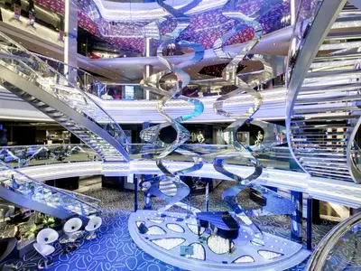 Das helle, moderne Infinity Atrium der MSC Grandiosa erstreckt sich über mehrere Ebenen mit glänzenden Treppen, einer großen Skulptur und blauer Beleuchtung.