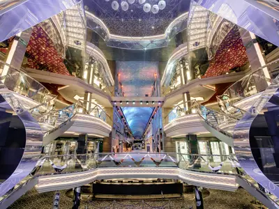 Das beeindruckende Infinity Atrium an Bord der MSC Grandiosa. Geschwungene Treppen, spiegelnde Oberflächen und eine digitale Decke mit Sternenhimmel. Geschäfte säumen die Promenade.