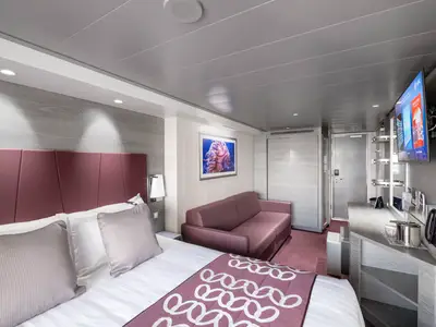 Balkonkabine an Bord der MSC Grandiosa. Zu sehen sind ein Doppelbett, ein Sofa, ein Schreibtisch und ein Fernseher. Moderne Einrichtung in Grau- und Weinrot-Tönen.