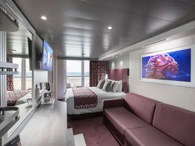 Moderne Balkonkabine an Bord der MSC Grandiosa mit Doppelbett, Sofa, TV und Balkon mit Meerblick. Ein großes Kunstwerk einer leuchtenden Qualle ziert die Wand über dem Sofa.