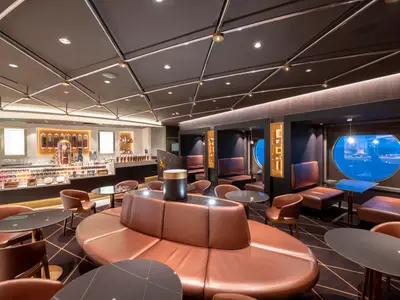 Eine moderne Bar und Lounge an Bord der MSC Grandiosa. Ledersitze, Holztische und ein Tresen mit Schokolade und Kaffee. Rundfenster bieten Ausblick.