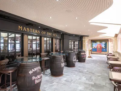 Die Bar 'Masters Of The Sea' an Bord der MSC Grandiosa. Dunkle Holzfassade, goldene Schrift, Fass-Tische. Im Gang rechts London-Wandmalerei mit roter Telefonzelle.