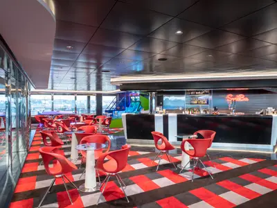 Eine lebhafte Sports Bar an Bord der MSC Grandiosa mit schwarzen und roten Akzenten. Moderne rote Stühle und Tische stehen auf einem gemusterten Teppich. Im Hintergrund sind Arkadespiele und eine Bar mit Neon-Schriftzug zu sehen. Große Fenster bieten einen Blick auf den Hafen.