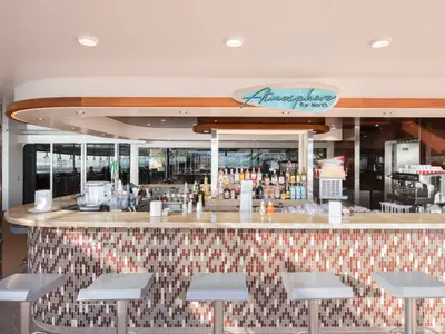 Blick auf die moderne Atmosphere Bar North an Bord der MSC Grandiosa mit gefliester Theke, ausgestellten Spirituosen, Kaffeemaschine und Barhockern. Im Hintergrund sind Fenster mit Aussicht zu sehen.