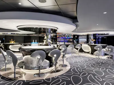 Blick auf die moderne Infinity Bar an Bord der MSC Grandiosa. Dunkle, geschwungene Bartheken mit integrierter Beleuchtung und elegante, graue Barhocker prägen das Design. Im Hintergrund sind Regale mit Spirituosen blau beleuchtet. Der Boden zeigt ein abstraktes Muster.