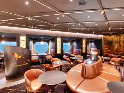 Innenansicht des Jean-Philippe Chocolat & Café an Bord der MSC Grandiosa. Warmer Raum mit Ledersitzen, Tischen und Bullaugen mit Meerblick. Goldene Wanddeko.