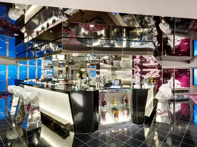 Blick in die schicke Champagne Bar an Bord der MSC Grandiosa. Eine lange, glänzende Theke mit geometrischen Hockern und leuchtenden Details ist zu sehen. Im Hintergrund sind blaue Fenster und lilafarbene Neonlichter mit der Aufschrift „CHAMPAGNE BAR“.