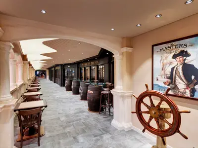 Langer Blick auf die maritime Promenade an Bord der MSC Grandiosa mit Säulenreihe links und dem Masters of the Sea Pub rechts. Im Vordergrund rechts ist ein hölzernes Schiffssteuerrad zu sehen, dahinter ein Gemälde eines Seefahrers.