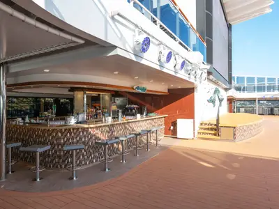 Aussenbar 'Atmosphere Bar North' auf dem Sonnendeck der MSC Grandiosa. Geschwungene Bar mit Mosaikfliesen und Barhockern. Eine Holzbuehne ist rechts daneben. Blauer Himmel und das Meer im Hintergrund.