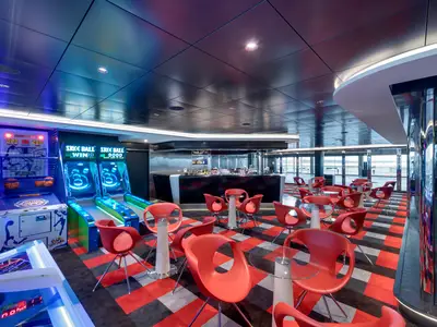 Die lebhafte Sports Bar an Bord der MSC Grandiosa. Zu sehen sind Arcade-Spiele, eine moderne Bar und rote Sessel mit Glastischen auf einem markanten schwarz-rot-grauen Teppich.