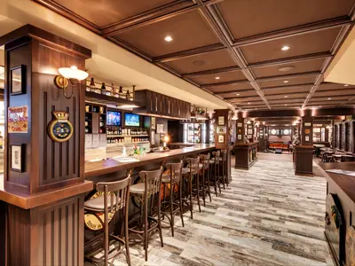 Eine lange, holzvertäfelte Bar-Lounge 'Masters of the Sea' an Bord der MSC Grandiosa mit Barhockern, dunklen Holzmöbeln und Flachbildschirmen über der Theke. An den Wänden sind maritime Dekorationen und Wandleuchten angebracht. Der Boden ist mit hellen Holzplanken ausgelegt, die Decke mit dunklen Kassetten verziert.