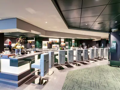 Edge Cocktail Bar an Bord der MSC Grandiosa. Lange, beleuchtete Theke mit weißen Barhockern. Regale voller Spirituosenflaschen im Hintergrund. Eleganter Innenbereich.