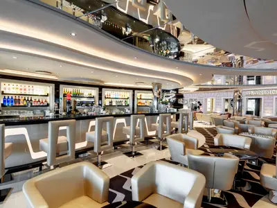 Elegante Grandiosa Bar & Lounge an Bord der MSC Grandiosa. Eine lange, beleuchtete Bar mit Barhockern und Flaschen. Im Hintergrund Sessel, Tische und ein Shop. Keine Personen sichtbar.