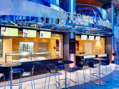 Ein moderner Jean-Philippe Crêpes & Gelato-Stand an Bord der MSC Grandiosa. Der Bereich ist in warmen Gelbtönen und kühlem Blau gehalten, mit Bildschirmen, Theken und dunklen Tischen und Stühlen.