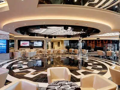 Blick in die elegante Grandiosa Bar & Lounge an Bord der MSC Grandiosa mit beigen Sesseln, Tischen, einer Bühne mit Instrumenten und einem auffälligen schwarz-weißen Bodenmuster.