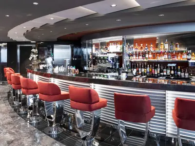 Elegante Bar-Lounge an Bord der MSC Grandiosa mit geschwungener Theke, roten Samt-Barhockern und einer grossen Auswahl an Spirituosen. Der Raum ist modern beleuchtet und hat einen polierten Marmorboden.