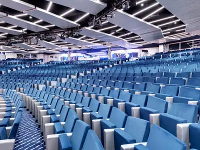 Das leere Théâtre La Comédie an Bord der MSC Grandiosa zeigt Reihen blauer Samtsitze. Moderne Deckenbeleuchtung und eine hell erleuchtete Bühne sind sichtbar.