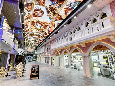 Die helle Galleria Grandiosa an Bord der MSC Grandiosa, eine Indoor-Promenade mit Geschäften, Restaurants und einer LED-Decke, die ein Deckengemälde zeigt. Ein Bistro mit Sitzgelegenheiten ist links sichtbar.