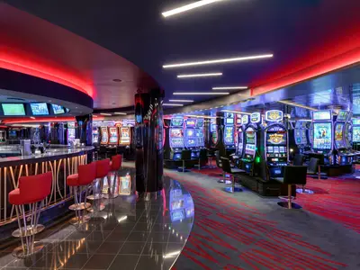 Modernes Casino an Bord der MSC Grandiosa. Es zeigt Reihen von beleuchteten Spielautomaten, eine stilvolle Bar mit roten Hockern und rote Deckenbeleuchtung.