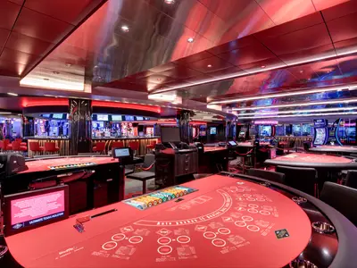Ein modernes Casino auf der MSC Grandiosa. Rote Spieltische, Spielautomaten und eine Bar sind in rotes Neonlicht getaucht. Der elegante Raum ist menschenleer.