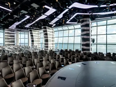 Eine helle, moderne Carousel Lounge an Bord der MSC Grandiosa, die viele Reihen dunkler Sitze und eine bühnenartige Fläche zeigt. Grosse Fenster bieten einen weiten Blick auf das offene Meer.