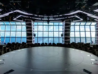 Die moderne Carousel Lounge an Bord der MSC Grandiosa bietet eine runde Bühne und grosse Panoramafenster mit Blick auf das offene Meer. Dunkle Sitzreihen umgeben die zentrale Bühne.