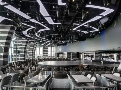 Die moderne Carousel Lounge an Bord der MSC Grandiosa. Eine Rundbühne, umgeben von Sitzreihen. An der Decke stilvolle Lichtleisten und Scheinwerfer. Seitlich große Fensterfronten.