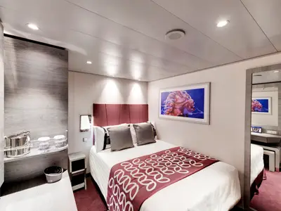 Innenansicht einer modernen Kabine an Bord der MSC Grandiosa. Ein Doppelbett mit weisser Bettwäsche und dunkelrotem Überwurf steht im Zentrum. An der Wand hängt ein farbenfrohes Kunstwerk.