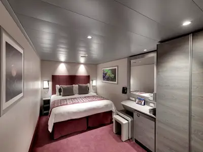 Ansicht einer modernen Innenkabine an Bord der MSC Grandiosa. Ein großes Doppelbett mit burgunderfarbenem Kopfteil und Teppich dominiert den Raum. Rechts befinden sich ein Schreibtisch mit Spiegel und ein kleiner Kühlschrank. Links ist ein gerahmtes Kunstwerk an der Wand.