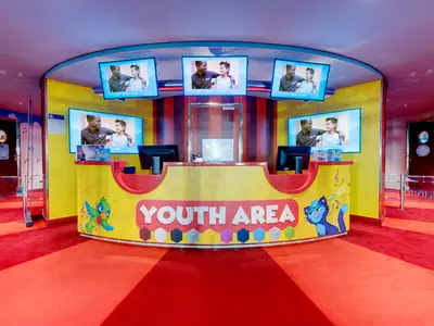 Bunter Jugendbereich an Bord der MSC Grandiosa. Eine Rezeption mit "YOUTH AREA"-Schriftzug, Bildschirmen. Roter Teppich und Türen zu Spielzimmern.