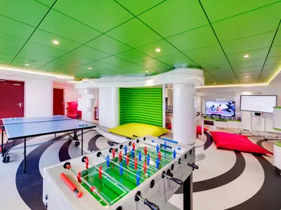 Ein lebhafter Spielebereich an Bord der MSC Grandiosa mit leuchtend grüner Decke, einer blauen Tischtennisplatte, einem Kicker, bequemen Sitzsäcken und Bildschirmen für Videospiele.