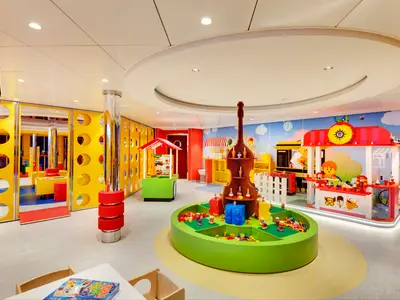 Ein farbenfroher LEGO Mini Club an Bord der MSC Grandiosa, ausgestattet mit verschiedenen Themen-Spielstationen, einer runden Baugrube voller LEGO-Steine und kinderfreundlichen Möbeln, die zum Spielen einladen. Die Wände sind mit einer Wolken- und Stadt-Wandmalerei versehen.