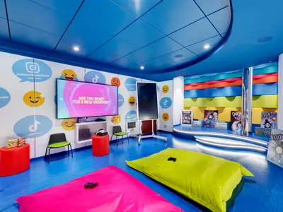 Der Web Creator Room an Bord der MSC Grandiosa zeigt einen lebhaften Raum mit blauer Einrichtung, großen Bildschirmen für Spiele und Social-Media-Wanddekorationen. Bunte Sitzkissen und Hocker laden zum Verweilen ein.