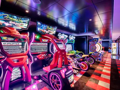 Eine moderne, neonbeleuchtete Spielhalle an Bord der MSC Grandiosa mit mehreren Arcade-Rennspielen, darunter 'Crusin' Blast' und Motorrad-Simulatoren. Der Boden hat ein rot-weiß kariertes Muster, das an eine Rennstrecke erinnert.
