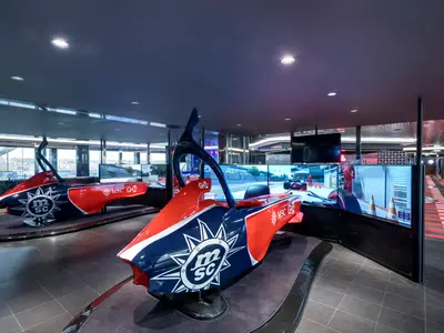 Zwei rote und blaue Formel-1-Rennsimulatoren mit MSC-Logo in einem hellen Unterhaltungsbereich an Bord der MSC Grandiosa. Grosse Bildschirme zeigen Rennspiel-Szenen. Moderne und sportliche Atmosphäre.