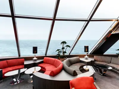 Blick in die stilvolle Top Sail Lounge der MSC Grandiosa. Grosse Panoramafenster bieten weiten Meerblick. Bequeme, geschwungene Sofas in Rot und Grau umrahmen Tische mit modernen Lampen.