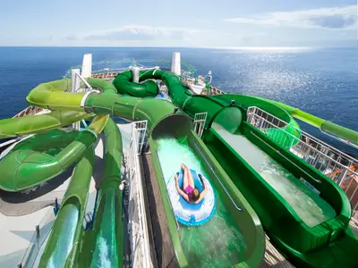 Eine Person auf blauem Reifen in einer grünen Wasserrutsche des Wild Forest Aquaparks der MSC Grandiosa. Grüne Röhrenrutschen schlängeln sich auf dem Deck. Im Hintergrund glitzert das weite Meer.