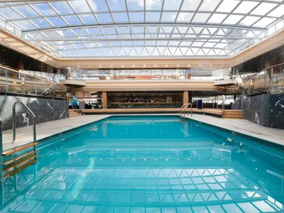 Ein geräumiger Poolbereich an Bord der MSC Grandiosa, mit grossem Schwimmbecken, eleganter Bar, dunklen Marmorwänden und transparentem Glasdach für viel Tageslicht. Ohne Personen.