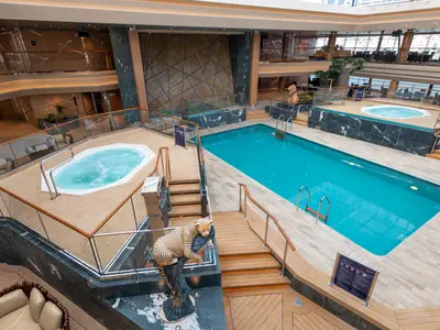 Safari Pool-Bereich der MSC Grandiosa. Großer Pool, zwei Whirlpools und eine Leoparden-Skulptur in luxuriösem Innenambiente mit Holz- und Marmorelementen.
