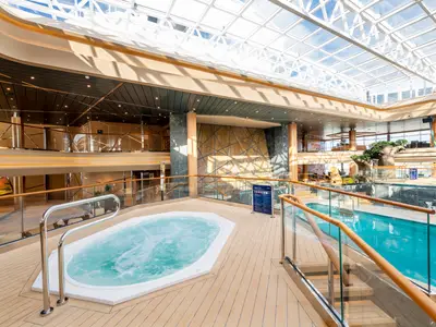 Der helle, mehrstöckige Indoor-Poolbereich an Bord der MSC Grandiosa. Ein sprudelnder Whirlpool und ein langer Pool, umgeben von Holzdecks und Glasgeländern unter einem weitläufigen Glasdach.