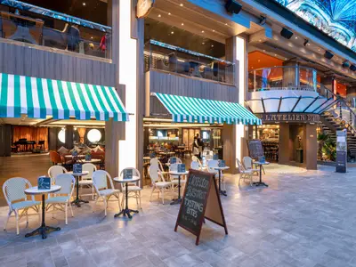 Eine helle, moderne Indoor-Promenade der MSC Grandiosa zeigt das L'Atelier Bistrot mit gestreiften Markisen und Aussenbestuhlung. Eine Skulptur und eine geschwungene Treppe sind sichtbar.